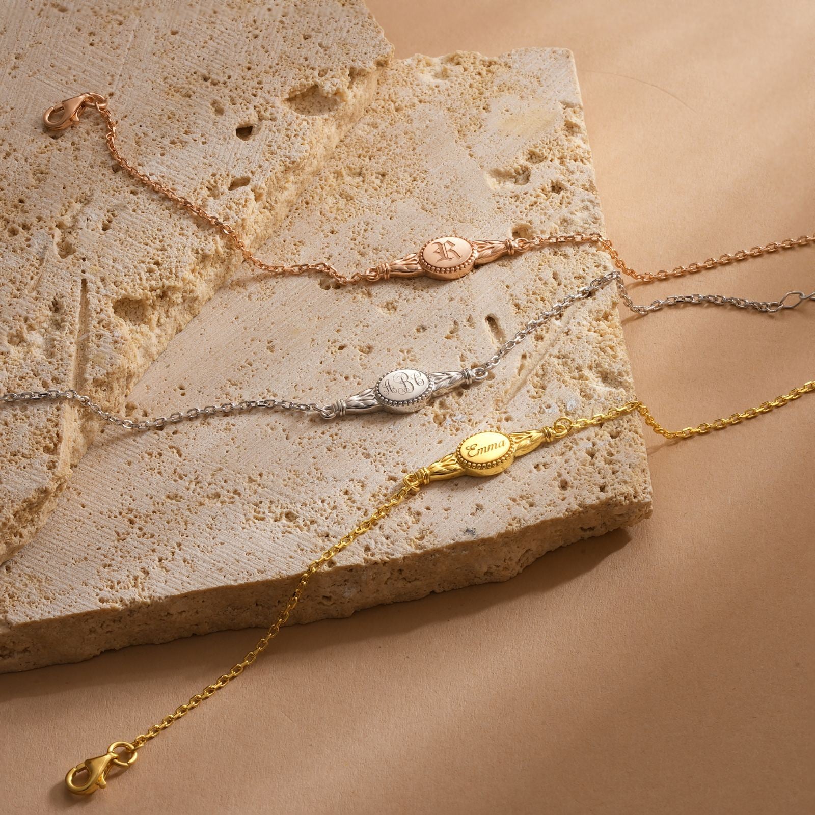 Engraved Oval Name Bracelet in three finishes: rose gold, silver, and yellow gold on textured stone background.