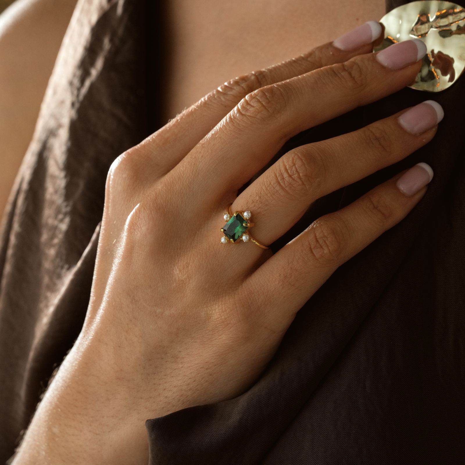 Hand wearing a gold Rectangle Statement Ring featuring a green birthstone and pearls against a brown fabric backdrop.